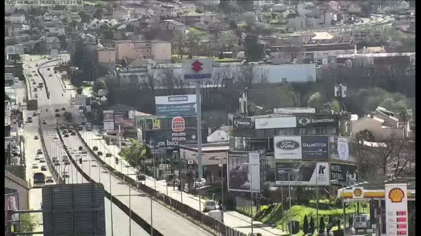A highway with moderate traffic flows through a city on a sunny day, passing numerous billboards and buildings.