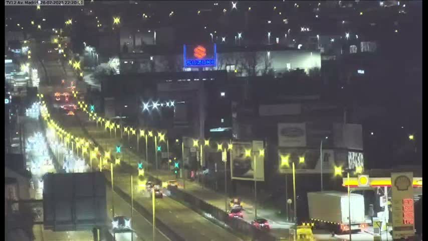 A nighttime, wet highway scene shows light traffic moving along a multi-lane roadway lined with illuminated streetlights, passing by various buildings and a gas station.