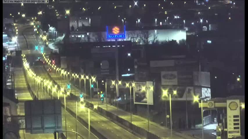 A nighttime, brightly lit highway scene shows several vehicles driving on a multi-lane road, passing under streetlights and alongside buildings with various advertisements.