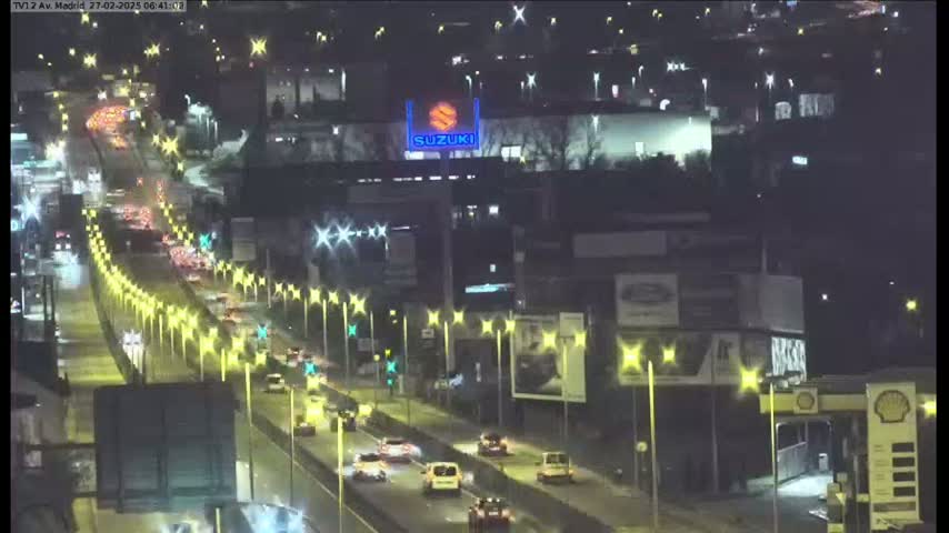 A nighttime view of a multi-lane highway with moderate traffic, illuminated by streetlights, passing by buildings and a gas station under a dark sky.
