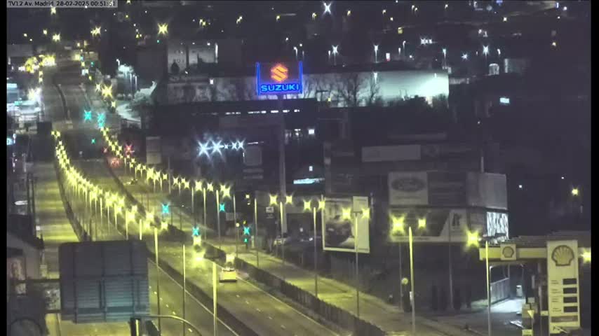 A brightly lit highway at night shows sparsely trafficked lanes lined with streetlights and buildings along the sides.