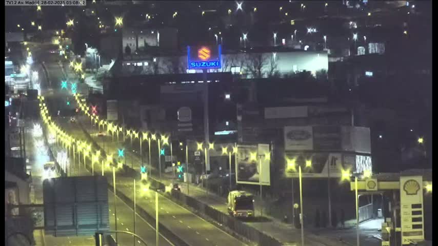A nighttime, brightly lit highway scene shows several vehicles traveling on a multi-lane road flanked by streetlights, buildings, and billboards.