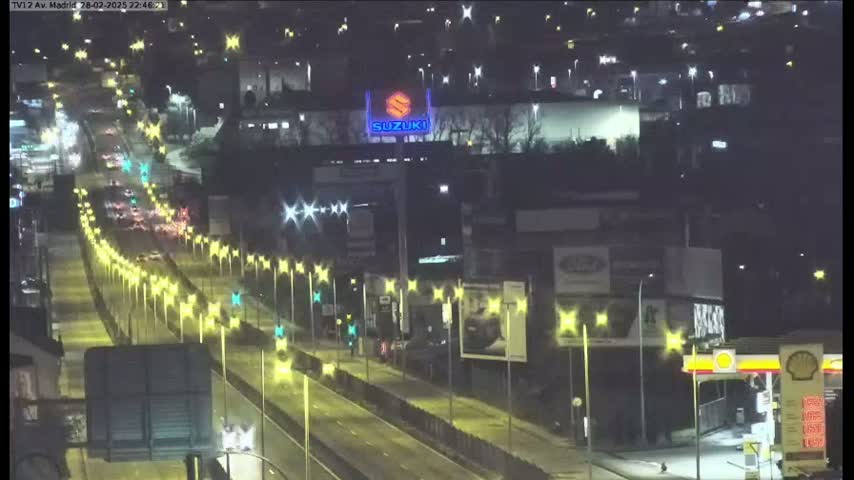 A nighttime, high-angle view of a multi-lane highway with moderate traffic, lined with streetlights and bordered by buildings and billboards.
