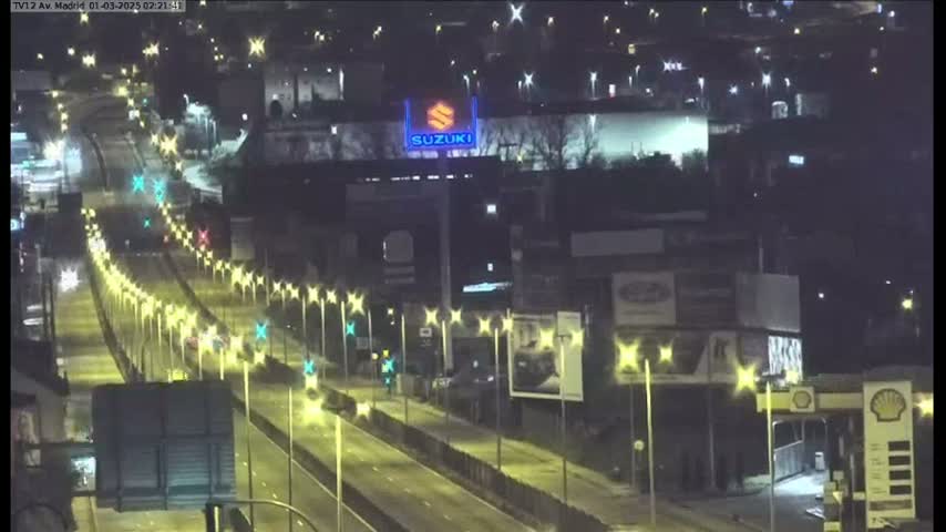 A nighttime, brightly lit highway scene shows numerous vehicles traveling along a multi-lane road flanked by streetlights and buildings.