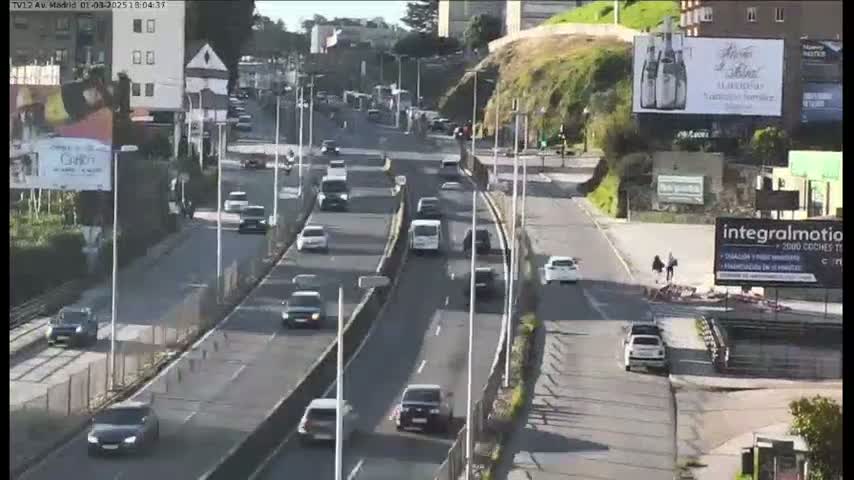 A multi-lane highway with moderate traffic flows under a sunny sky, with buildings and greenery visible on either side.