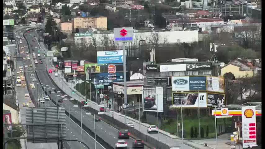A highway scene with numerous vehicles driving on a multi-lane road, surrounded by buildings and billboards under an overcast sky.