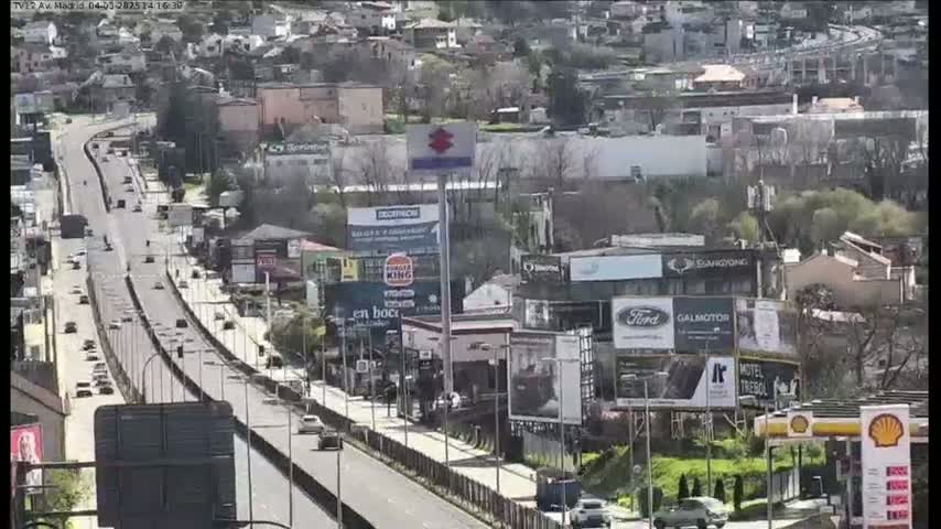 A highway with moderate traffic flows through a city on a sunny day, passing numerous billboards and buildings.