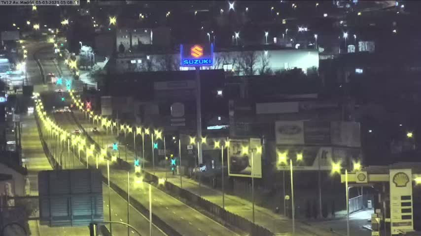 A nighttime, brightly lit highway with sparse traffic, bordered by buildings and billboards.