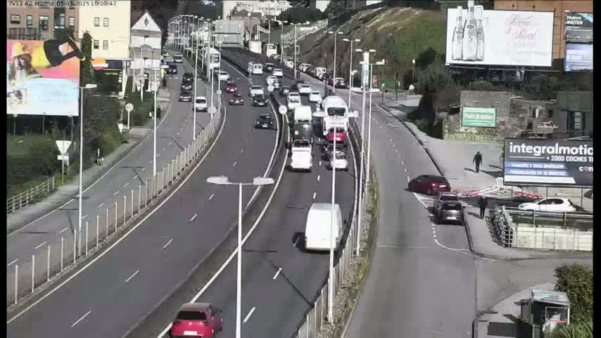A sunny day shows a multi-lane highway with moderate traffic flow, featuring several cars and trucks moving in both directions, alongside a pedestrian walkway and buildings.