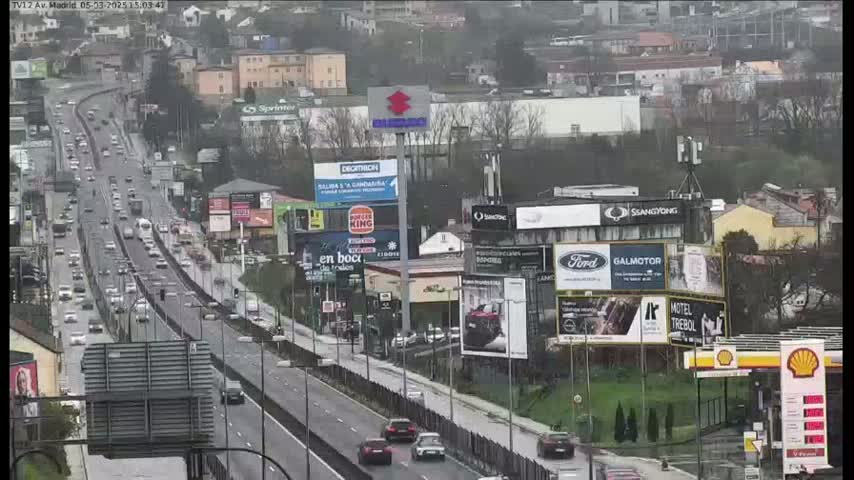 A highway with moderate traffic flows through a city under overcast skies, surrounded by billboards and buildings.