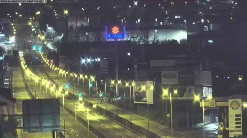 A nighttime, brightly lit highway scene shows a few cars traveling on a multi-lane road lined with streetlights, passing by buildings and billboards.