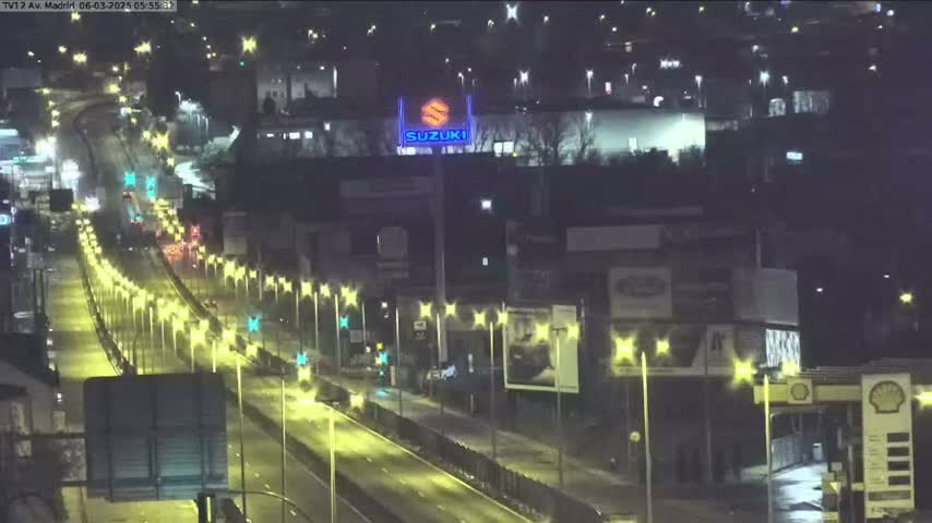 A nighttime, brightly lit highway scene shows several vehicles traveling along a multi-lane roadway flanked by streetlights and buildings, with a large Suzuki logo visible in the distance.
