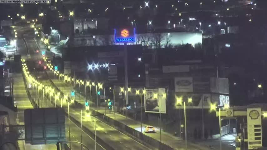 A brightly lit highway at night, with sparse traffic, runs through a city with buildings and advertisements visible along the sides.