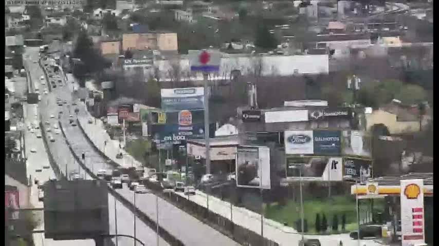 A highway with moderate traffic flows through a city, bordered by buildings and billboards under overcast skies.