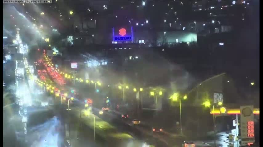 A rain-slicked highway at night shows heavy traffic flowing under bright streetlights, with a Suzuki logo visible in the distance.
