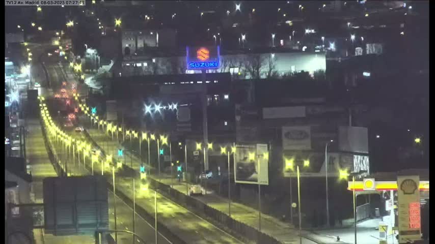 A nighttime, wet highway scene shows multiple vehicles traveling on a multi-lane road lined with illuminated streetlights, passing buildings and advertisements.