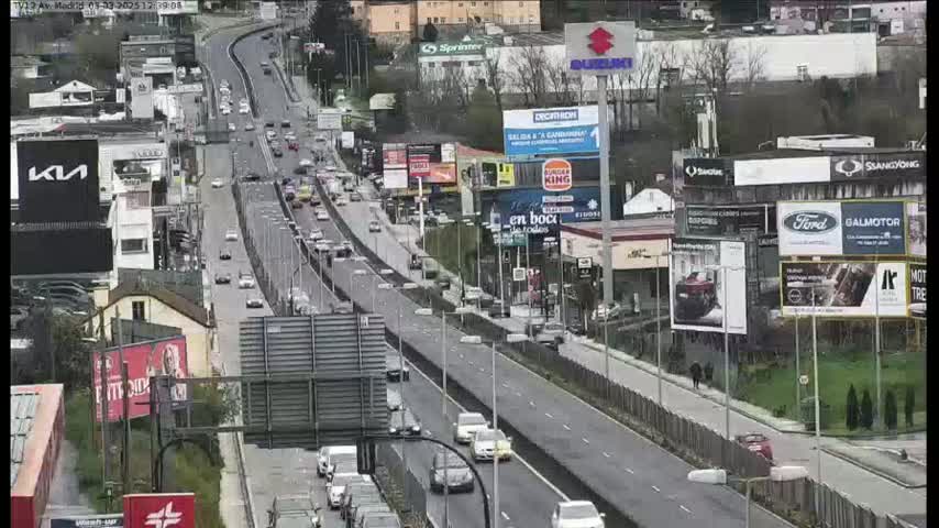 A highway scene on a partly cloudy day shows numerous vehicles traveling in both directions, passing by various businesses and billboards.