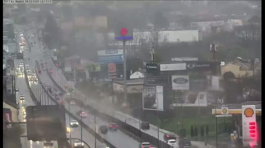 A rain-swept highway scene shows numerous vehicles driving in heavy rain past billboards and buildings.