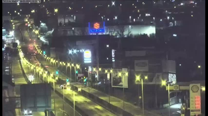 A nighttime, high-angle view of a multi-lane roadway with sparse traffic, lined with streetlights, and buildings alongside it.