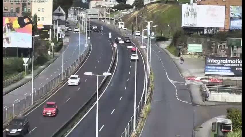 A multi-lane highway with several cars driving on a sunny day curves past buildings and billboards.
