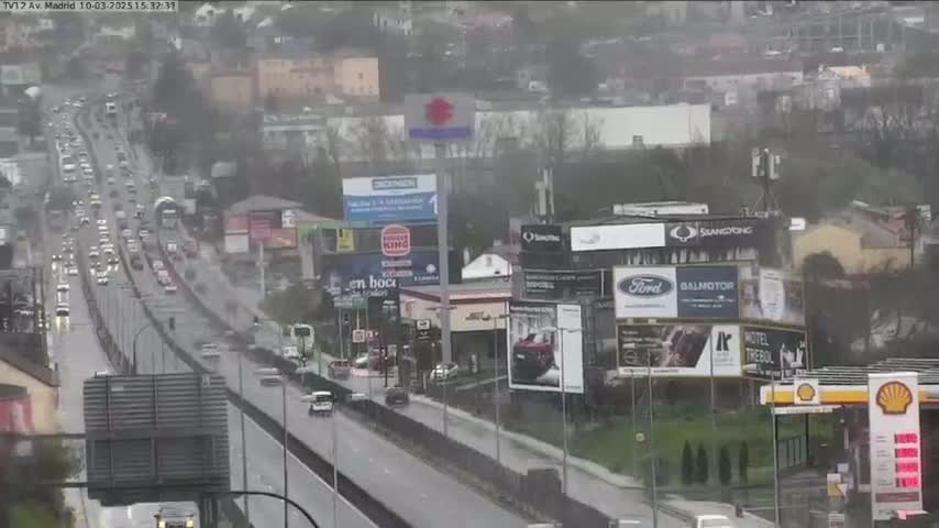 A rain-soaked highway with moderate traffic flows through a city with numerous billboards and buildings.