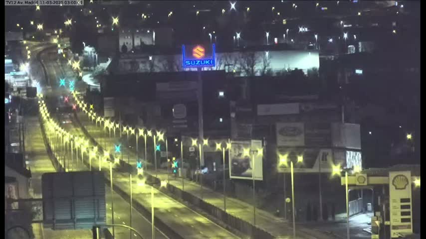 A nighttime, brightly lit highway with sparse traffic stretches into the distance, passing under a large Suzuki sign and beside a gas station.