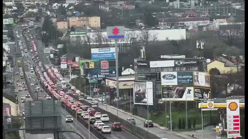 A long line of mostly red cars is stopped in heavy traffic on a multi-lane road on an overcast day, with numerous billboards and buildings visible in the background.