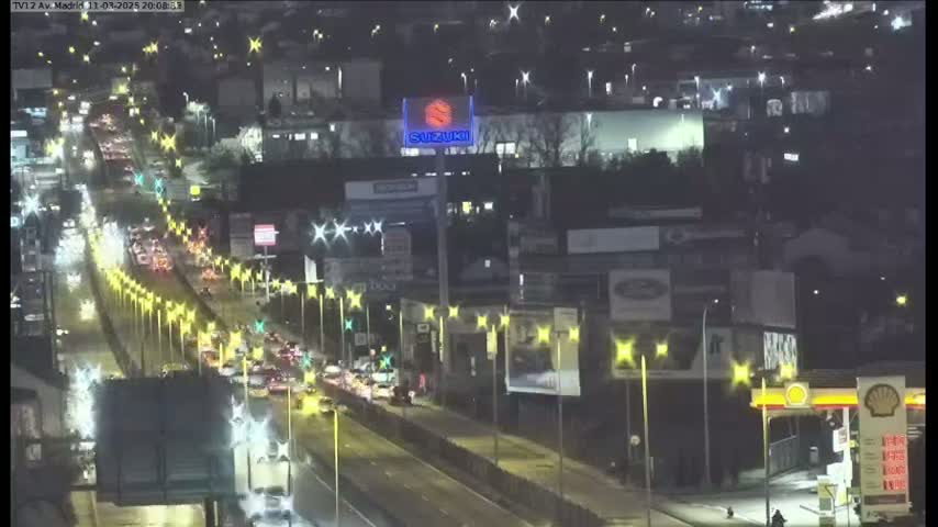 A nighttime, slightly overcast view of a multi-lane highway with moderate traffic flow, lined with illuminated streetlights and bordered by buildings and billboards.