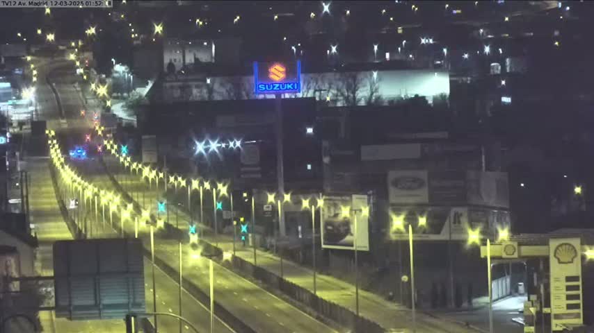 A nighttime, brightly lit highway scene shows several vehicles traveling along a multi-lane road lined with lampposts, passing by buildings and billboards.