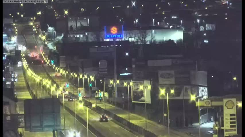 A nighttime, brightly lit highway scene shows several cars driving along a multi-lane road lined with illuminated streetlights, passing by various businesses and billboards.