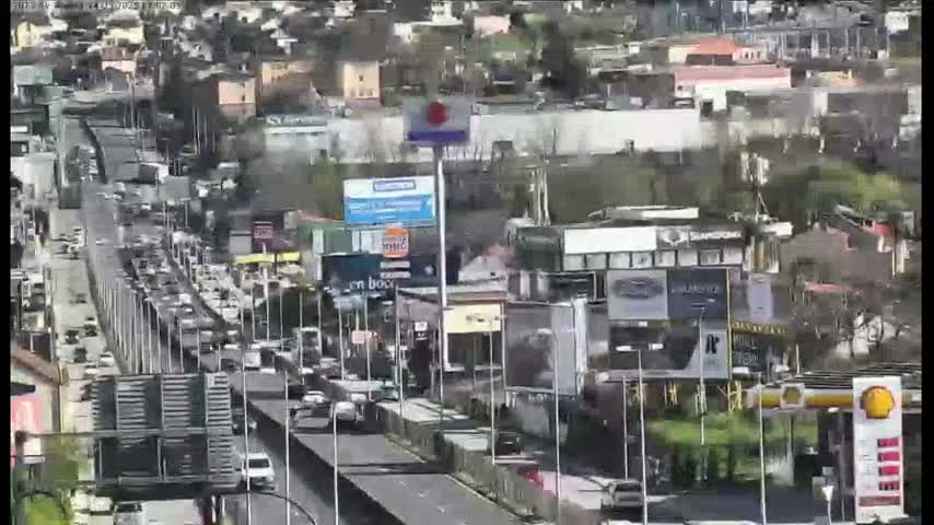 A busy highway with heavy traffic flows past buildings and businesses on a sunny day.