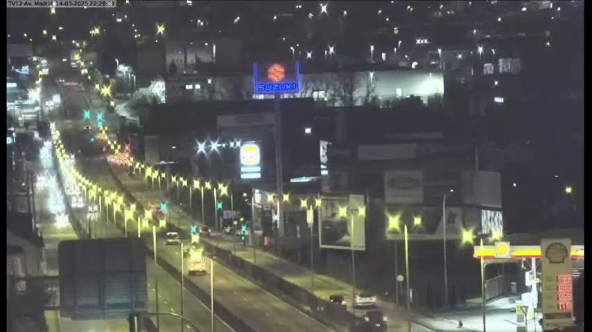 A nighttime view of a multi-lane highway with moderate traffic, lined with streetlights and buildings, including a large Suzuki advertisement.