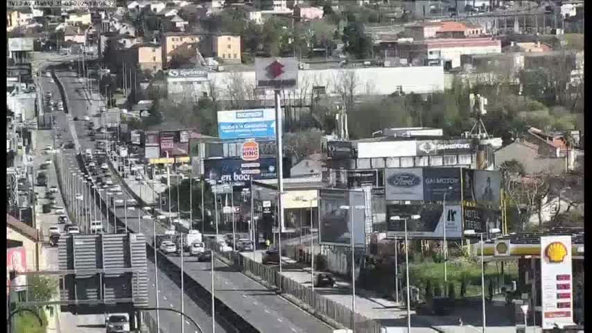 A highway with heavy traffic flows through a city on a sunny day, surrounded by buildings and billboards.