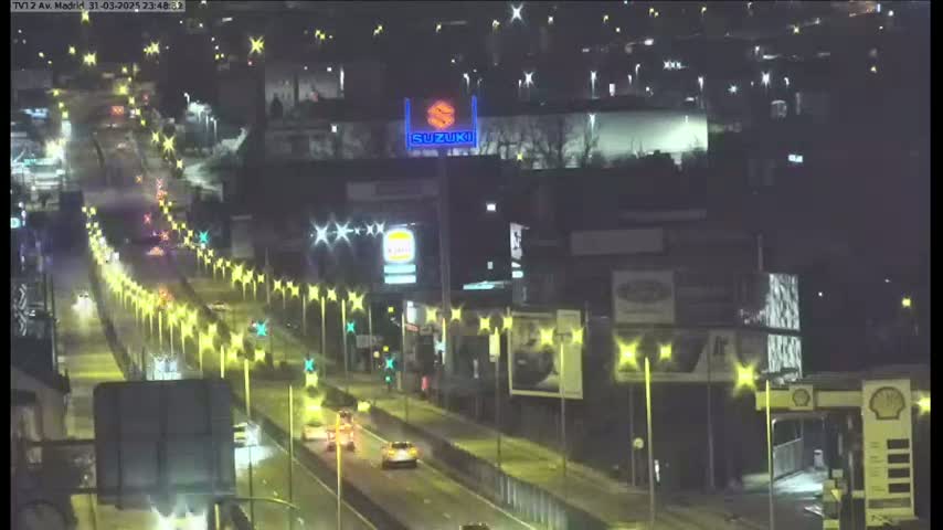 A nighttime view of a multi-lane highway with sparse traffic, lined with streetlights and buildings, including a gas station and a Suzuki advertisement.