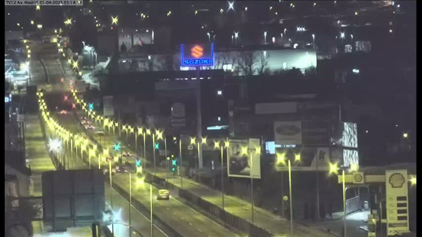 A nighttime, brightly lit highway scene shows several vehicles traveling along a multi-lane road lined with lights, passing by various buildings and billboards.