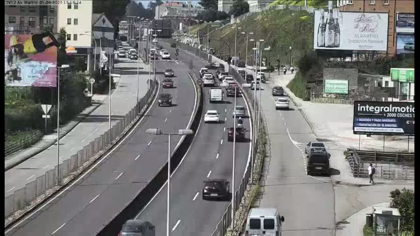 A multi-lane highway with moderate traffic flows under sunny conditions, surrounded by buildings and some greenery.
