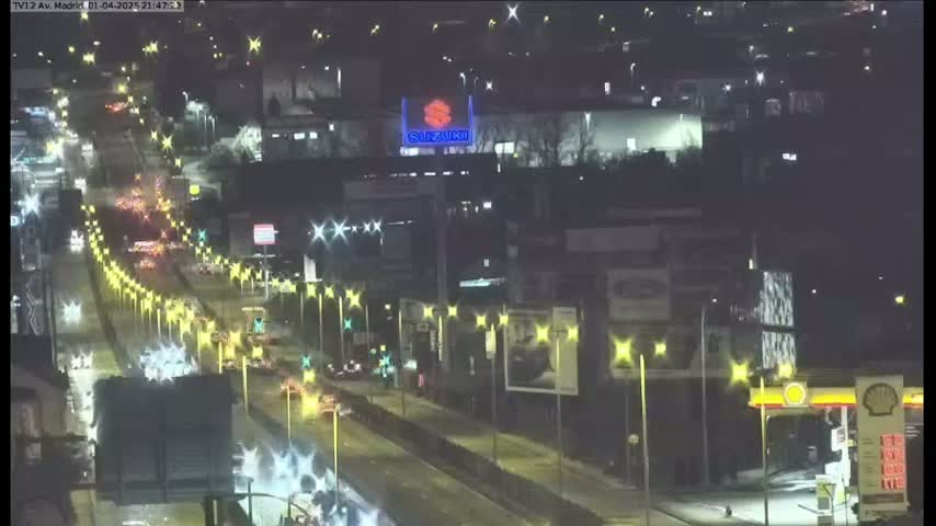 A nighttime, brightly lit highway with moderate traffic flows past buildings and businesses.