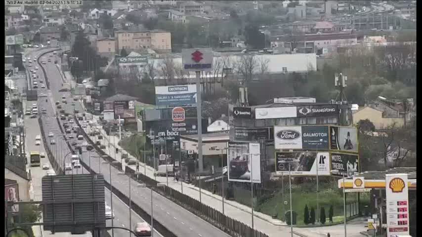 A highway with moderate traffic is seen, alongside a city with numerous billboards and buildings, under an overcast sky.