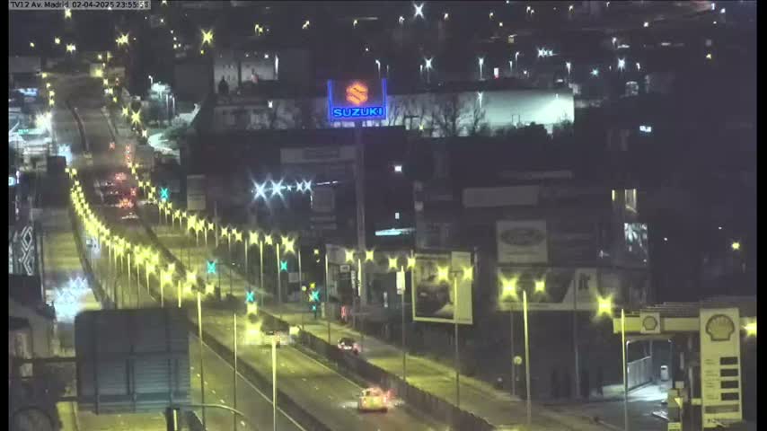 A nighttime, rain-slicked highway scene shows a multi-lane road with sparse traffic, lined with illuminated streetlights, passing under a large illuminated building.