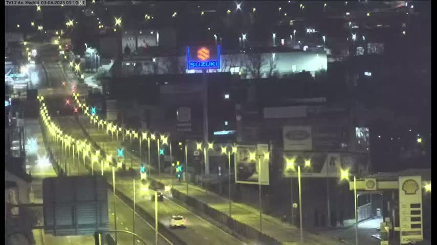 A wet, nighttime highway scene shows several vehicles driving on a multi-lane road lined with illuminated streetlights, passing in front of buildings and a Suzuki dealership sign.