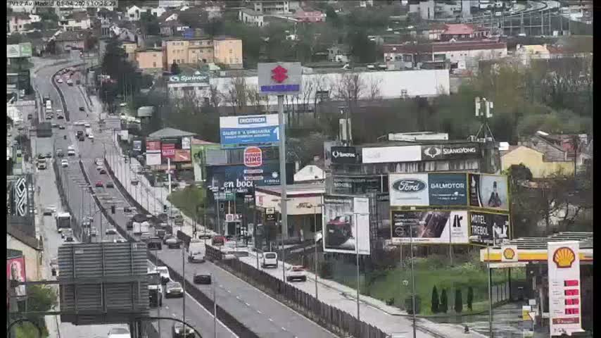 A highway with moderate traffic flows through a city with numerous buildings and billboards, under overcast skies.