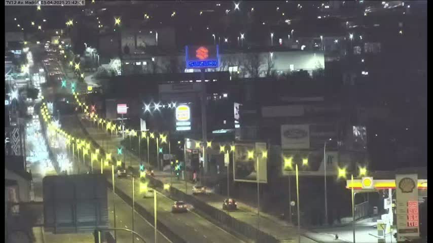 A nighttime, aerial view of a multi-lane highway with moderate traffic, lined with streetlights and buildings, including a gas station and a billboard.