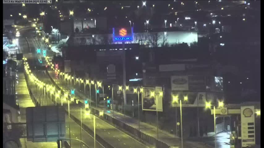 A nighttime, wet highway scene shows sparse traffic flowing under illuminated streetlights past commercial buildings and a large Suzuki sign.