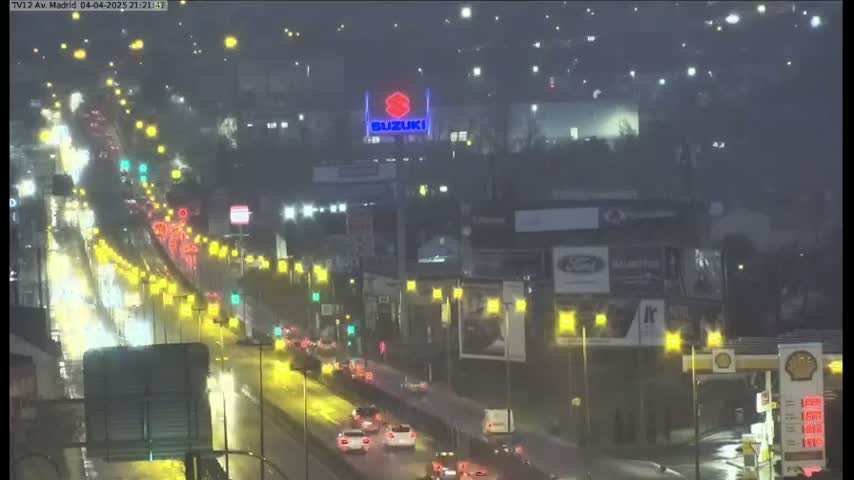 A rain-slicked highway at night is filled with headlights of slow-moving traffic, passing illuminated buildings and billboards.