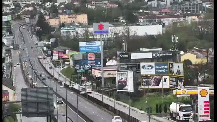 A highway with moderate traffic flows through a town, lined with billboards and businesses, under a mostly cloudy sky.