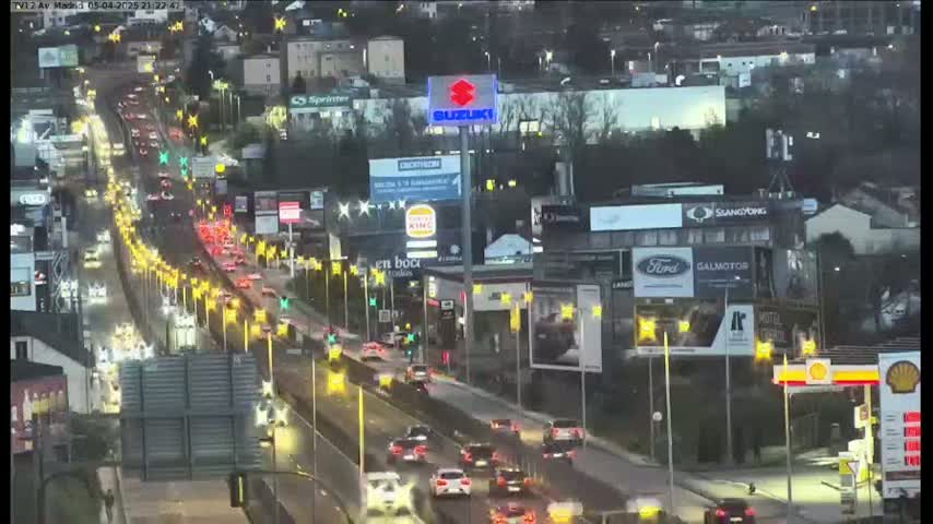 A nighttime, slightly dark aerial view of a busy highway with numerous cars and buildings with billboards illuminated along the sides.