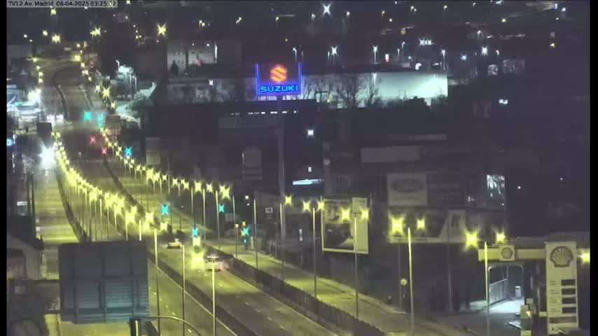 A nighttime, lightly trafficked highway stretches past a building with a Suzuki logo illuminated, all under a clear sky.
