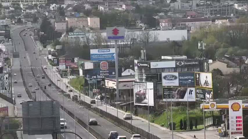 A highway scene on a partly cloudy day shows light traffic moving along a multi-lane road lined with billboards and buildings, including a gas station.