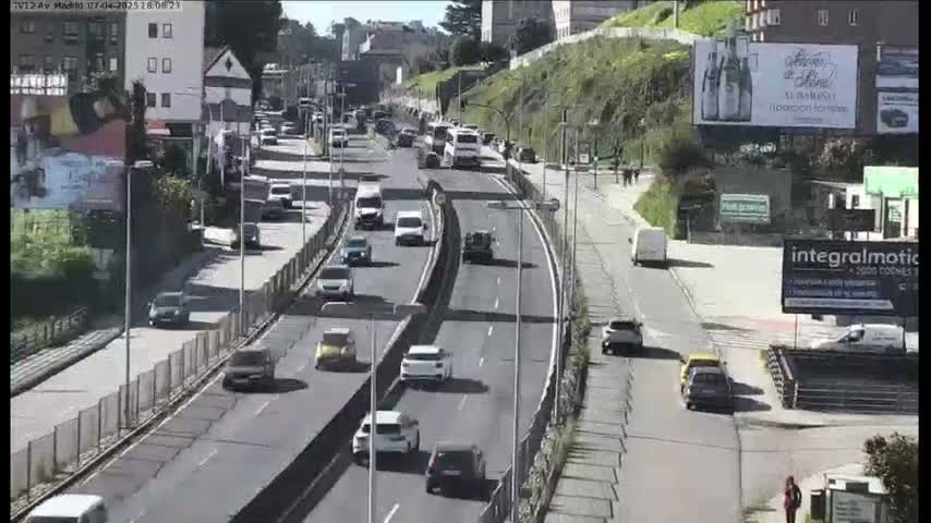 A sunny day shows a multi-lane highway with moderate traffic flow, featuring cars and buses navigating through a city with buildings and greenery visible on the sides.