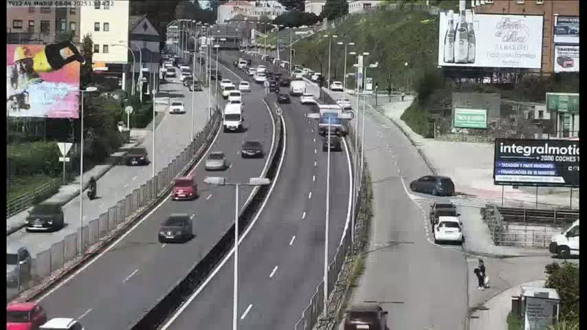 A sunny day shows a multi-lane highway with moderate traffic curving through a city, alongside a pedestrian walkway and buildings.